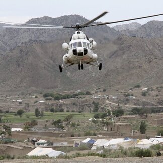 A white helicopter hovers above a rural area with tents and buildings. The background features mountainous terrain with scattered vegetation and structures.